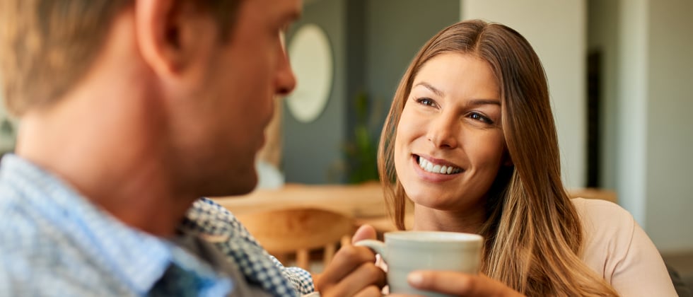 A woman and a man sitting next to each other. The woman has a cup in her hands and smiles at the guy.