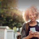 Black single woman smiles in her smartphone while dating online on the go in UK