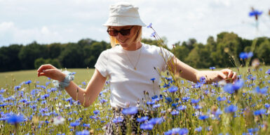 A woman standing on a field of violet flowers. She looks happy and is waring a hat and sunglasses.