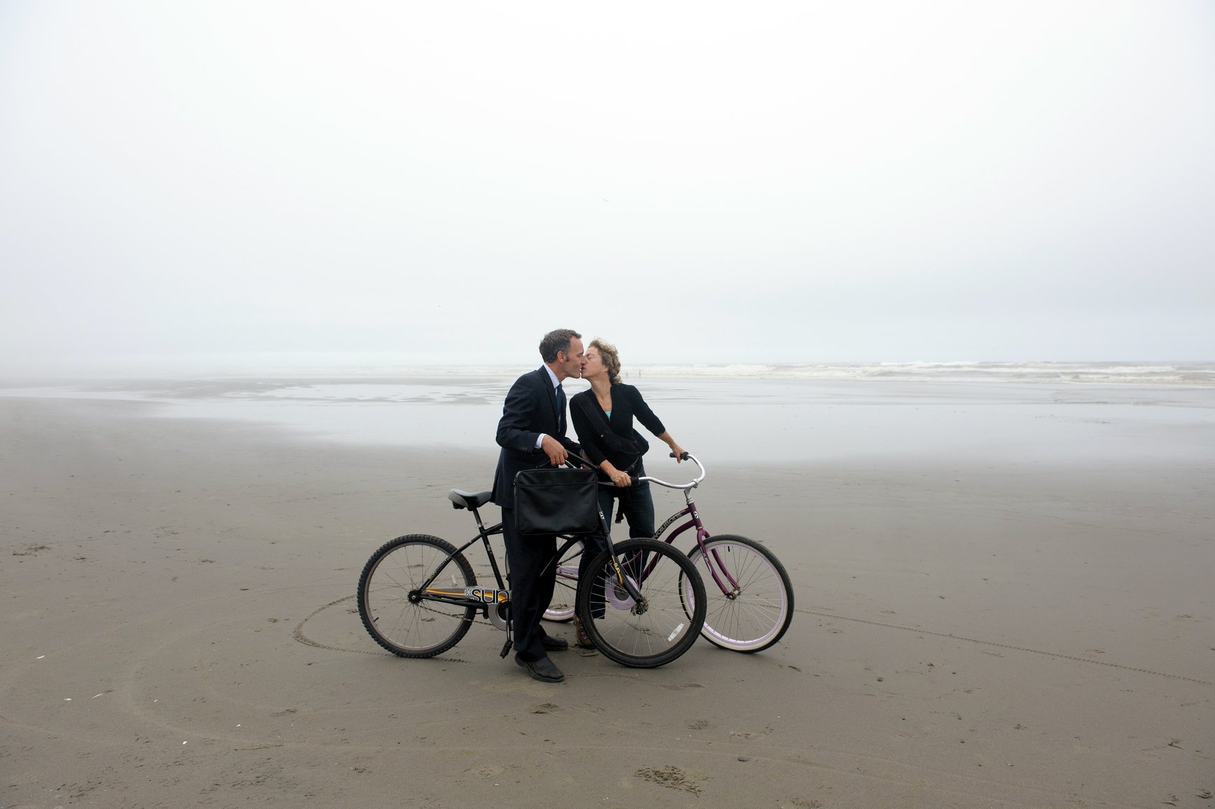 A man and woman sitting on bikes on a misty beach kissing each other. 