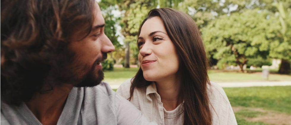 A man and a woman sitting next to each other in nature. She is looking at him happily and showing a love language.