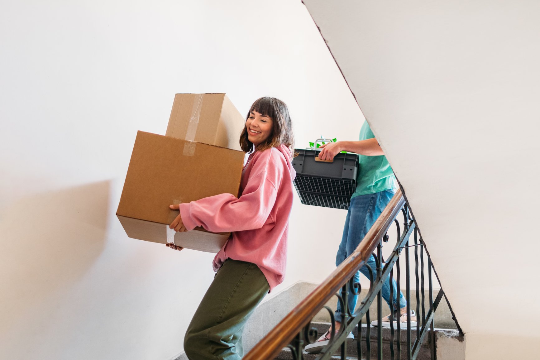 Young love couple walking down staircase, woman carrying cardboard boxes and man carrying crate with house plant and other stuff, moving in new building together